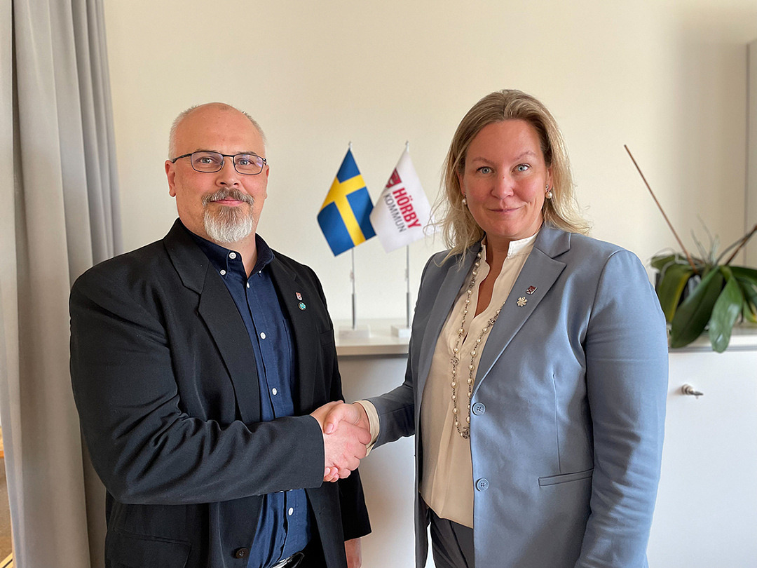 Anders Hansson och Cecilia Bladh in Zito skakar hand med Hörby kommunflagga och svenska flaggan i bakgrunden.