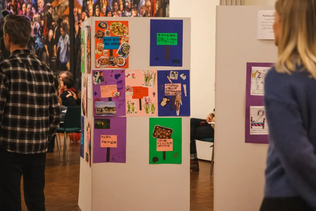Färgglada teckningar och kollage gjorda av barn är uppsatta på en vikvägg. Folk står runtomkring och kollar på dem
