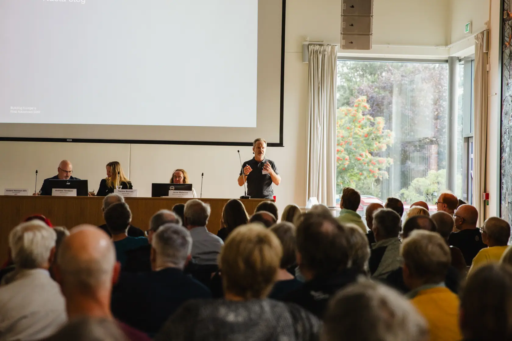 Ryggar på sittande publik som tittar mot ett presidiebord med sittande människor