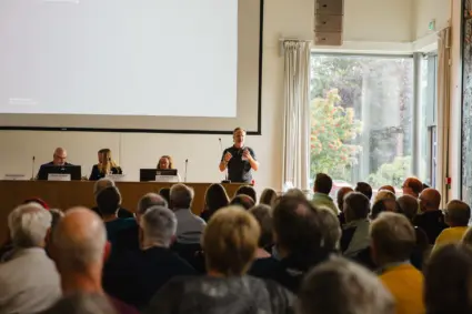 Ryggar på sittande publik som tittar mot ett presidiebord med sittande människor