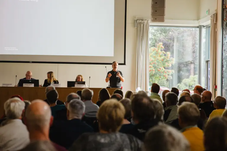 Ryggar på sittande publik som tittar mot ett presidiebord med sittande människor