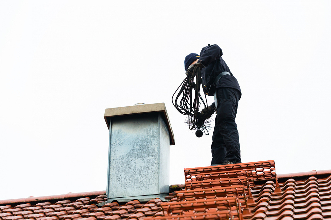 Svartklädd person med sotningsredskap på ett hustak intill en skorsten.