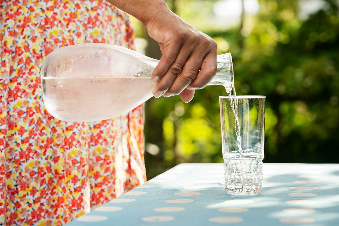 Med en glaskaraff i handen häller någon upp bubblande klart vatten i ett glas. Glaset står på ett bord i solen.