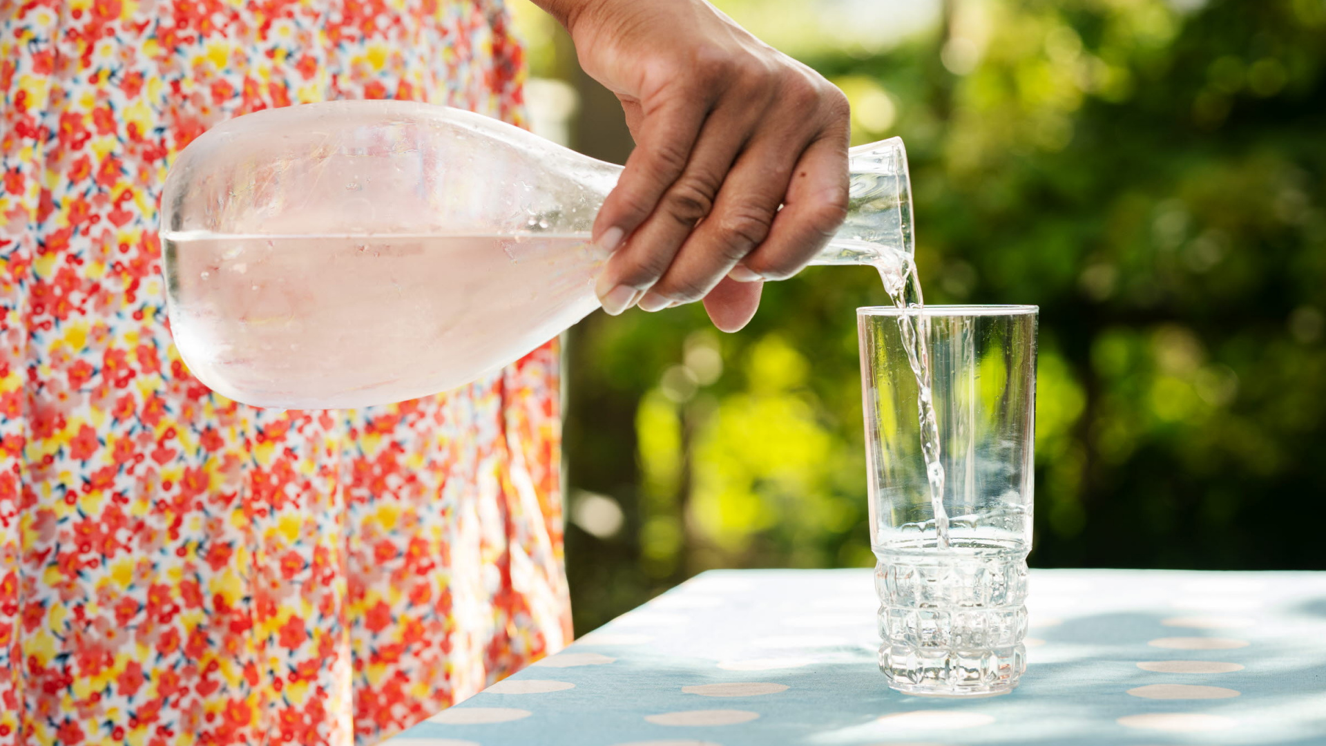 Med en glaskaraff i handen häller någon upp bubblande klart vatten i ett glas. Glaset står på ett bord i solen.
