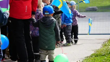 Närbild på ballong i ett barns hand, barnet går i ett led med andra barn och vuxna