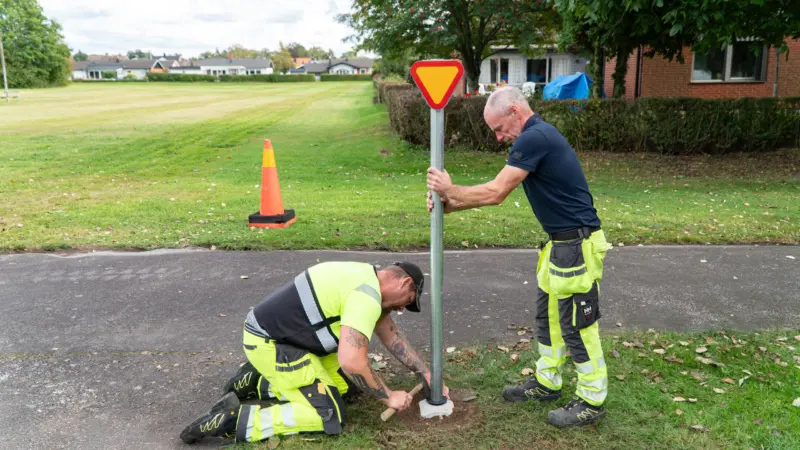 Två män monterar en stolpe med vägskylt i miniatyr i marken. Ena mannen lägger jord över fundamentet medan den andra mannen håller stolpen rak.