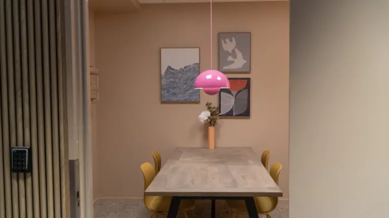 Mötesrum fotad utifrån, med mjukt persikofärgade väggar med tavlor på, rosa taklampa, träbord med stolar.