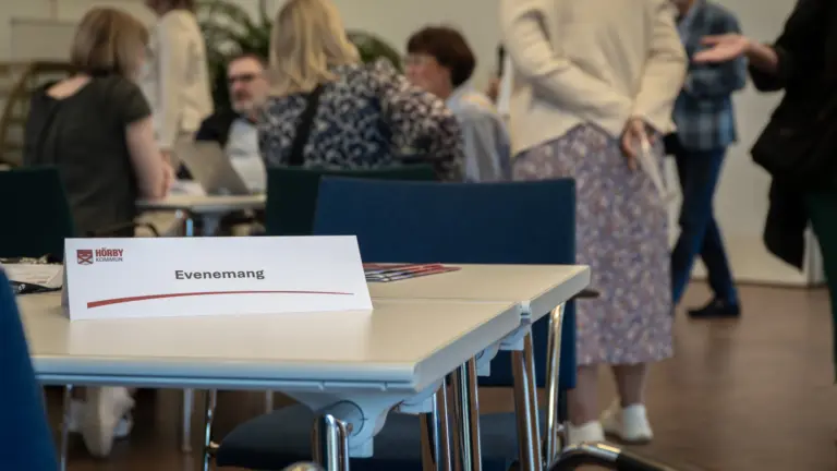 Samling människor i bakgrunden, bord med skylten "evenemang" i förgrunden.