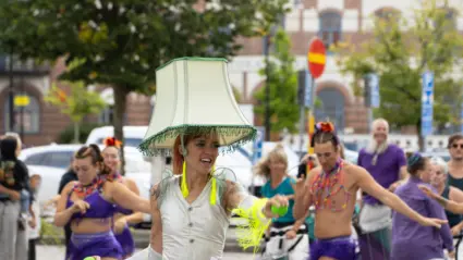 En grupp människor dansar och spelar trummor på en gata. I mitten syns en person med en stor vit lampskärm på huvudet.