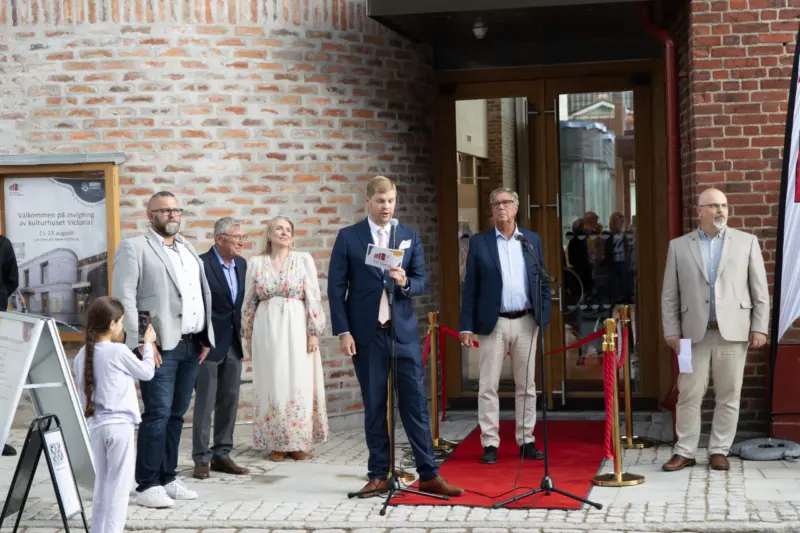 En grupp personer står utanför en tegelbyggnad vid en invigning. En man håller i en mikrofon och en röd matta ligger utanför entrén.