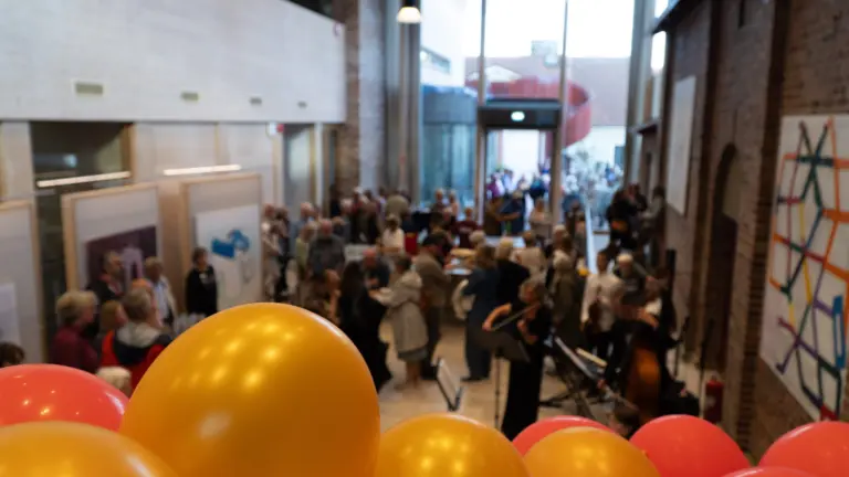 En sal full av människor som minglar. I förgrunden syns guldiga och röda ballonger.