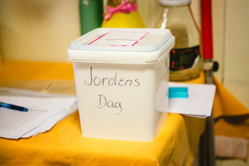 En vit plastbytta med texten "Jordens Dag" i svart tusch skriven på framsidan. Plastbyttan står på ett bord med gul duk. I bakgrunden syns diverse papper och en penna.