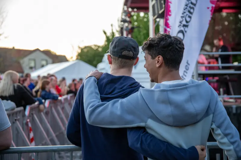 Tv&aring; killar fotograferade bakifr&aring;n st&aring;r med varsin arm vilandes p&aring; den andres axel. I bakgrunden syns en scen, kravallstaket och en suddig publik.