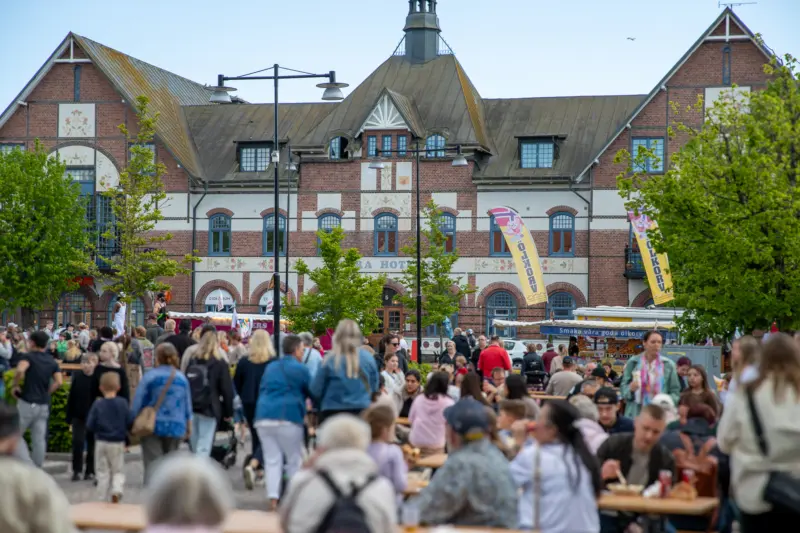 En stor folkmassa best&aring;ende av b&aring;de barn och vuxna sitter utspridda vid olika bord i kv&auml;llssolen p&aring; ett torg. I bakgrunden syns ocks&aring; f&auml;rgglada food trucks och framf&ouml;r dessa syns l&aring;nga k&ouml;er av m&auml;nniskor.