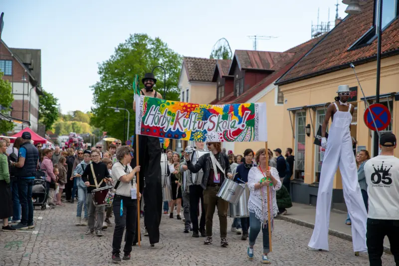 En grupp m&auml;nniskor g&aring;r under en f&auml;rgglad, handm&aring;lad banderoll d&auml;r det st&aring;r "H&ouml;rby Kulturskola!". Bilden &auml;r tagen utomhus.