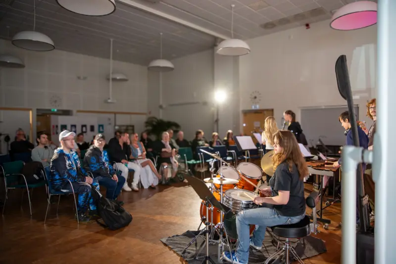 Ett band best&aring;ende av trummor, klarinetter, xylofon och gitarr spelar framf&ouml;r en publik i en stor, nedsl&auml;ckt sal. 