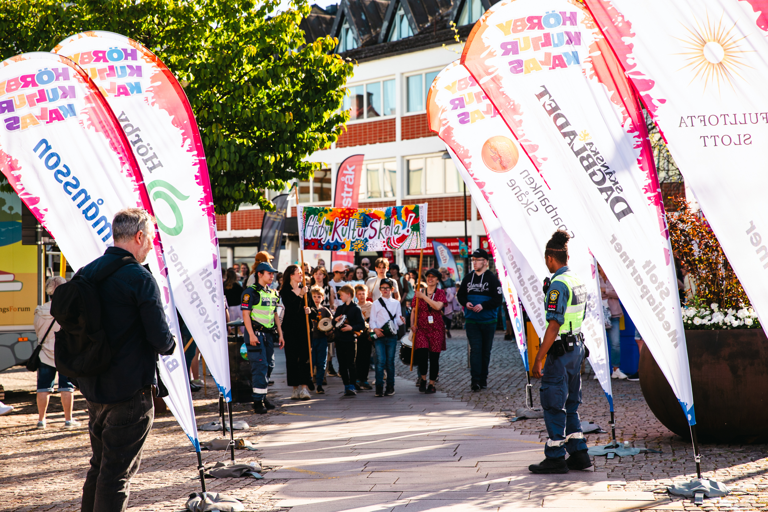 En grupp människor går under en färgglad, handmålad banderoll där det står "Hörby Kulturskola!". Bilden är tagen utomhus och i förgrunden syns några människor samt färgglada beachflaggor med Hörby Kulturkalas logotyp.