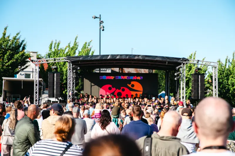 En bild på en scen fylld av ett 50-tal barn i 10-årsåldern. De blickar ut över publiken bestående av föräldrar, som har sina bakhuvuden vända mot kameran. Ovanför och bakom scenen syns en klarblå himmel och gröna träd.