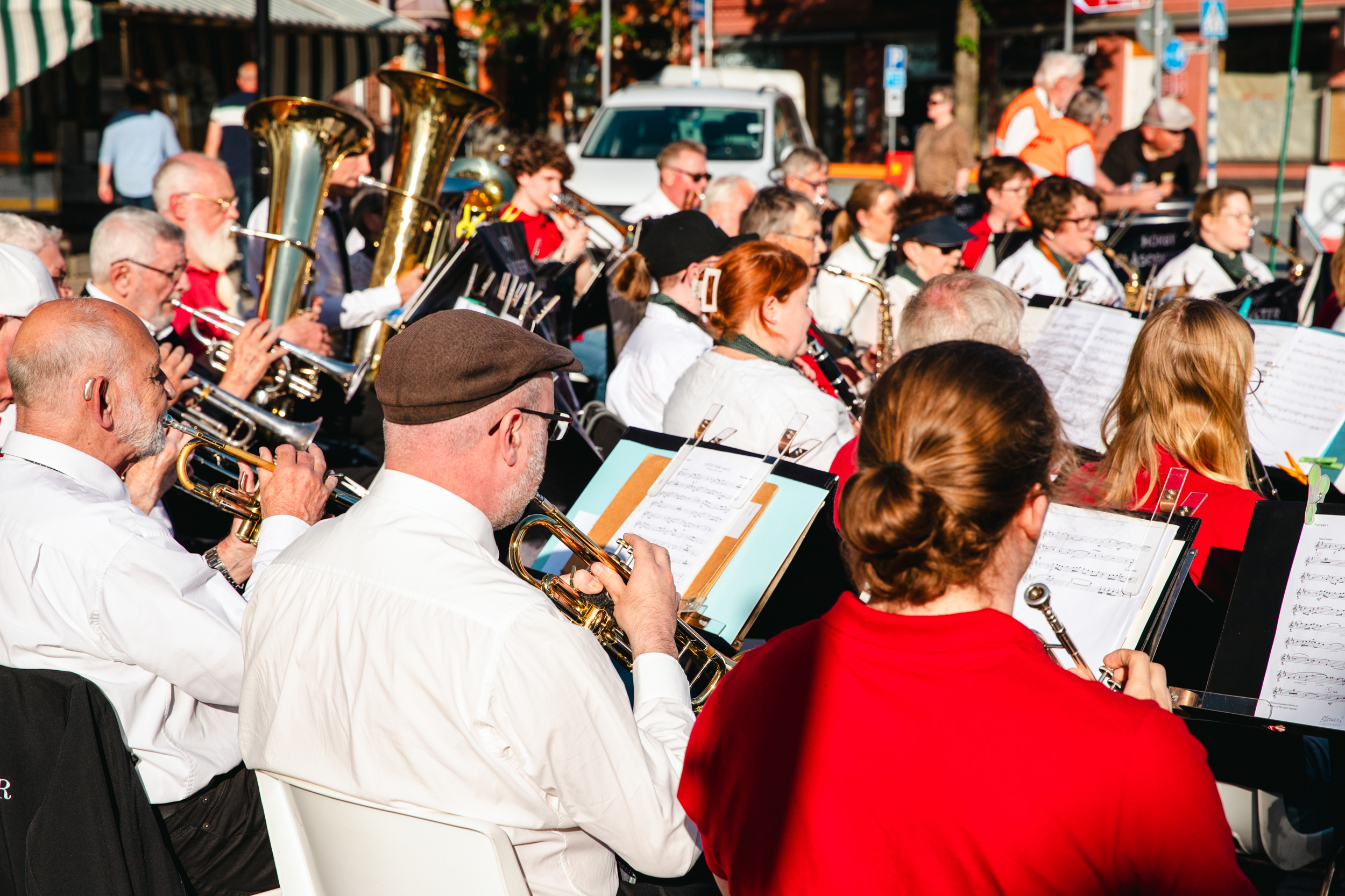 En orkester med blåsinstrument fotograferad bakifrån. Förutom musikanterna syns också deras notstativ och mässingsinstrument som blänker i solen.