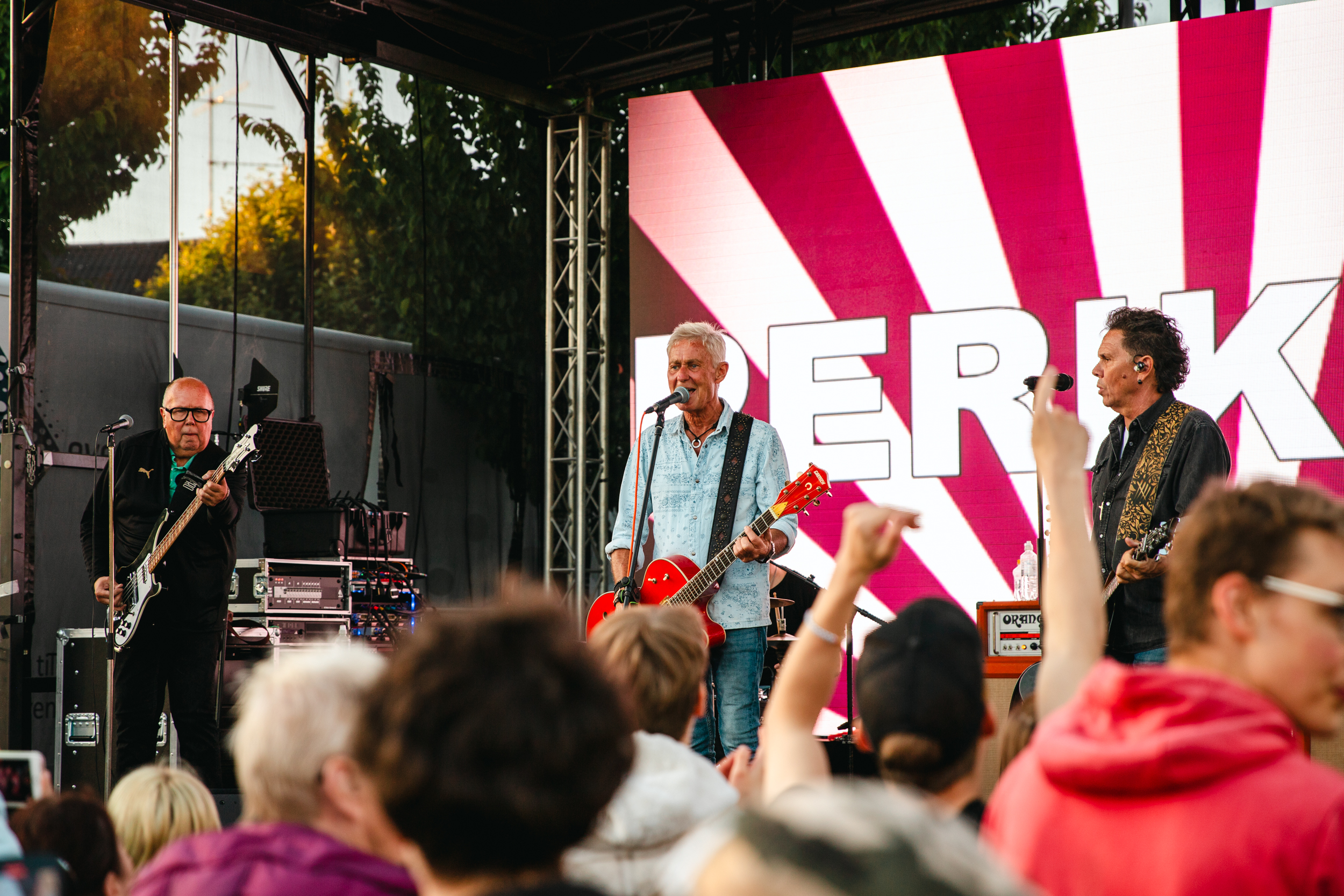 Tre män står på scen, med varsin gitarr i handen. Publiken i förgrunden tittar hängivet på musikanterna, en ung kille sträcker upp sina händer och armar i luften när han dansar med.