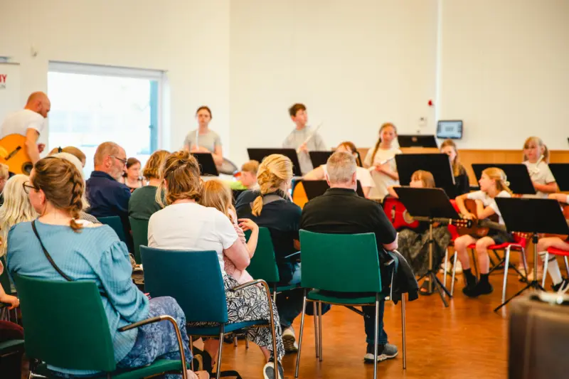 På bilden, som är tagen inomhus, sitter en publik framför en orkester av barn. Hälften av barn har en gitarr i famnen, resten står redo med sina tvärflöjtar.