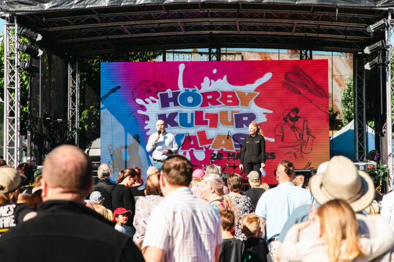 En man och en kvinna står på en scen och talar ut i mikrofon till publiken, som består av både barn och vuxna. På scenen syns också en färgglad LED-skärm med Hörby Kulturkalas logotyp