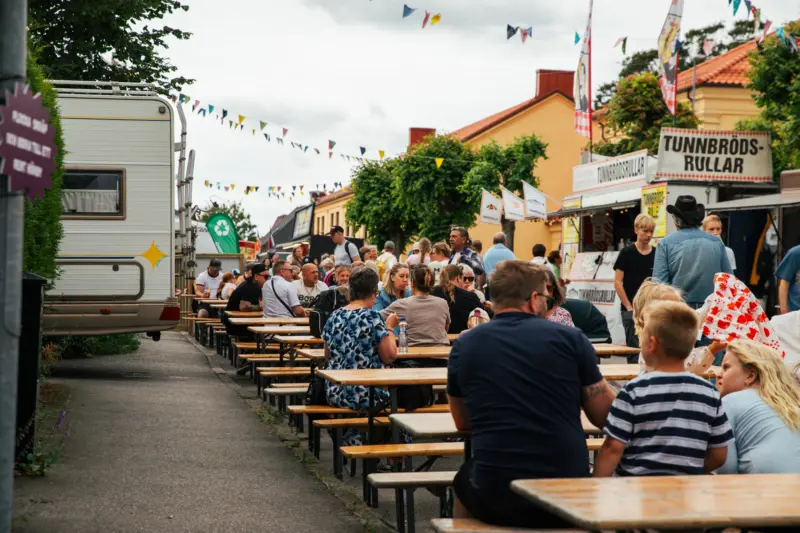Olika sällskap av personer sitter och äter vid uppställda bord på en asfalterad gata utomhus.