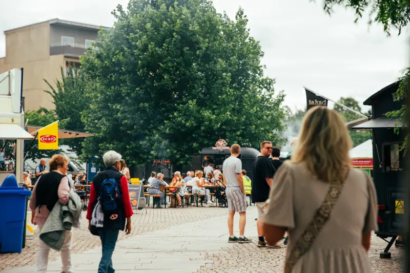 På ett torg med kullersten står många besökare som alla har ryggen mot kameran. I bakgrunden syns bord där folk sitter och äter, samt rök från en grill.