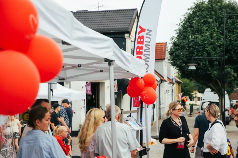 En handfull personer står under och utanför ett vitt partytält och samtalar med varandra. Det är en molnig dag och i förgrunden syns röda ballonger.