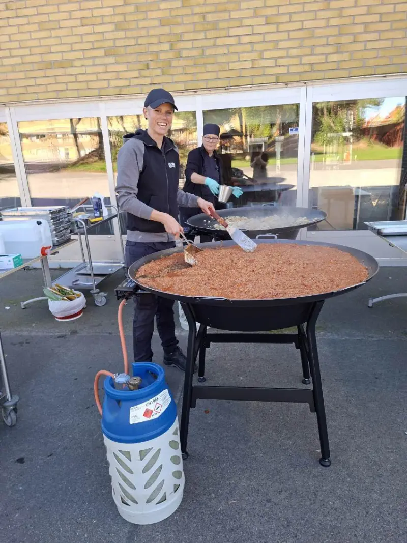 Bild tagen utomhus på två glada personer som står och lagar mat på en stor stekhäll.