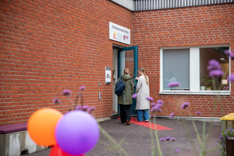 tv&aring; m&auml;nniskor som g&aring;r in genom en d&ouml;rr till en byggnad. I f&ouml;rgrunden syns ballonger och v&auml;xter.