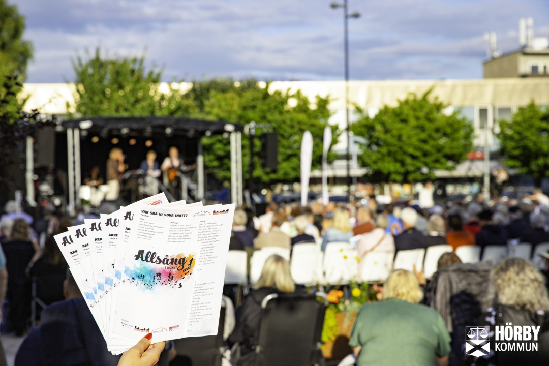 Hand håller ett gäng sånghäften till Allsång med en scen och fullsatt publik i bakgrunden