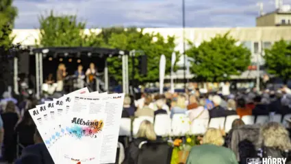Hand håller ett gäng sånghäften till Allsång med en scen och fullsatt publik i bakgrunden