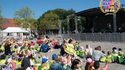 flera barn sitter på marken framför en scen och tittar på två människor som gör rörelser.