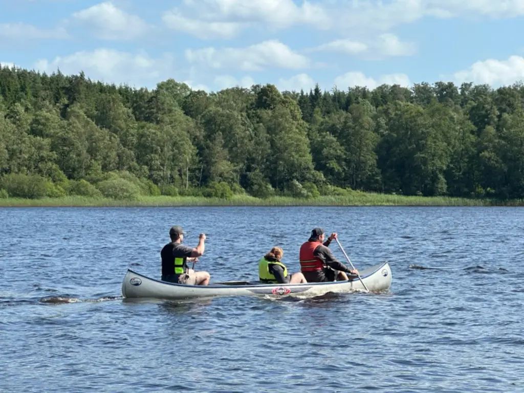 Tre personer sitter i en kanot och paddlar bort från kameran. Vattnet och himlen är blått, och på andra sidan vattendraget syns ett grönt skogsbryn.