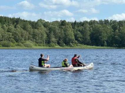 Tre personer sitter i en kanot och paddlar bort från kameran. Vattnet och himlen är blått, och på andra sidan vattendraget syns ett grönt skogsbryn.
