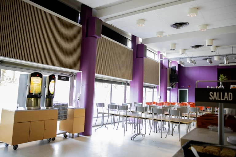 Skolrestaurang med stora fönster, lila väggar och ljusa träpaneler. Rader med bord och prydligt uppsatta stolar.