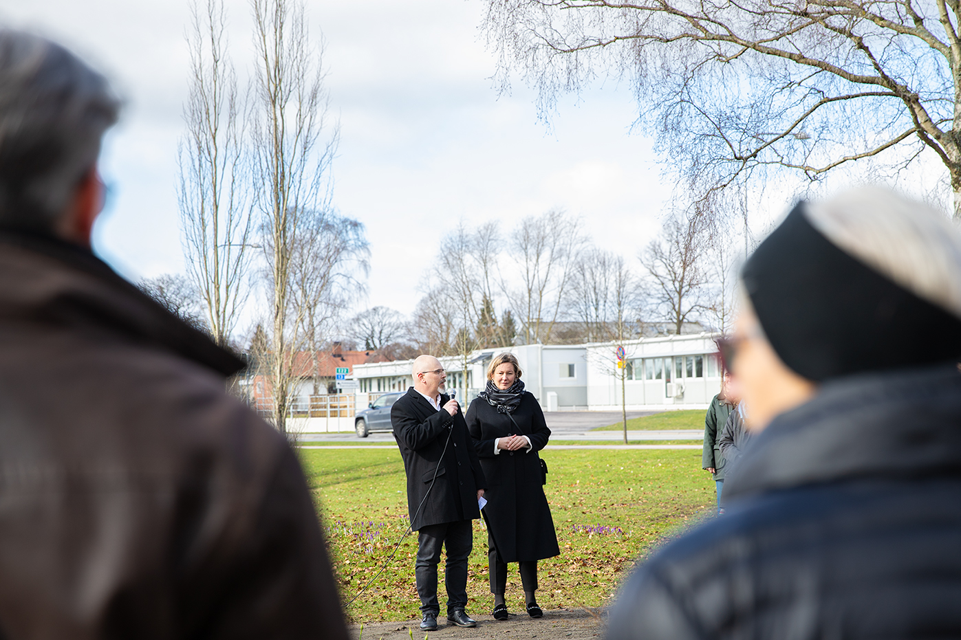 En man som står med mikrofon och en kvinna står utomhus och håller ett tal till åskådare. 