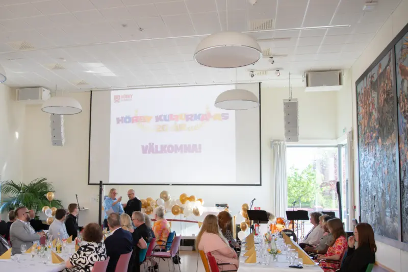 uppdukade långbord fullt med folk. Längst fram står två personer och håller tal. En projektor projicerar kulturkalas-logotypen