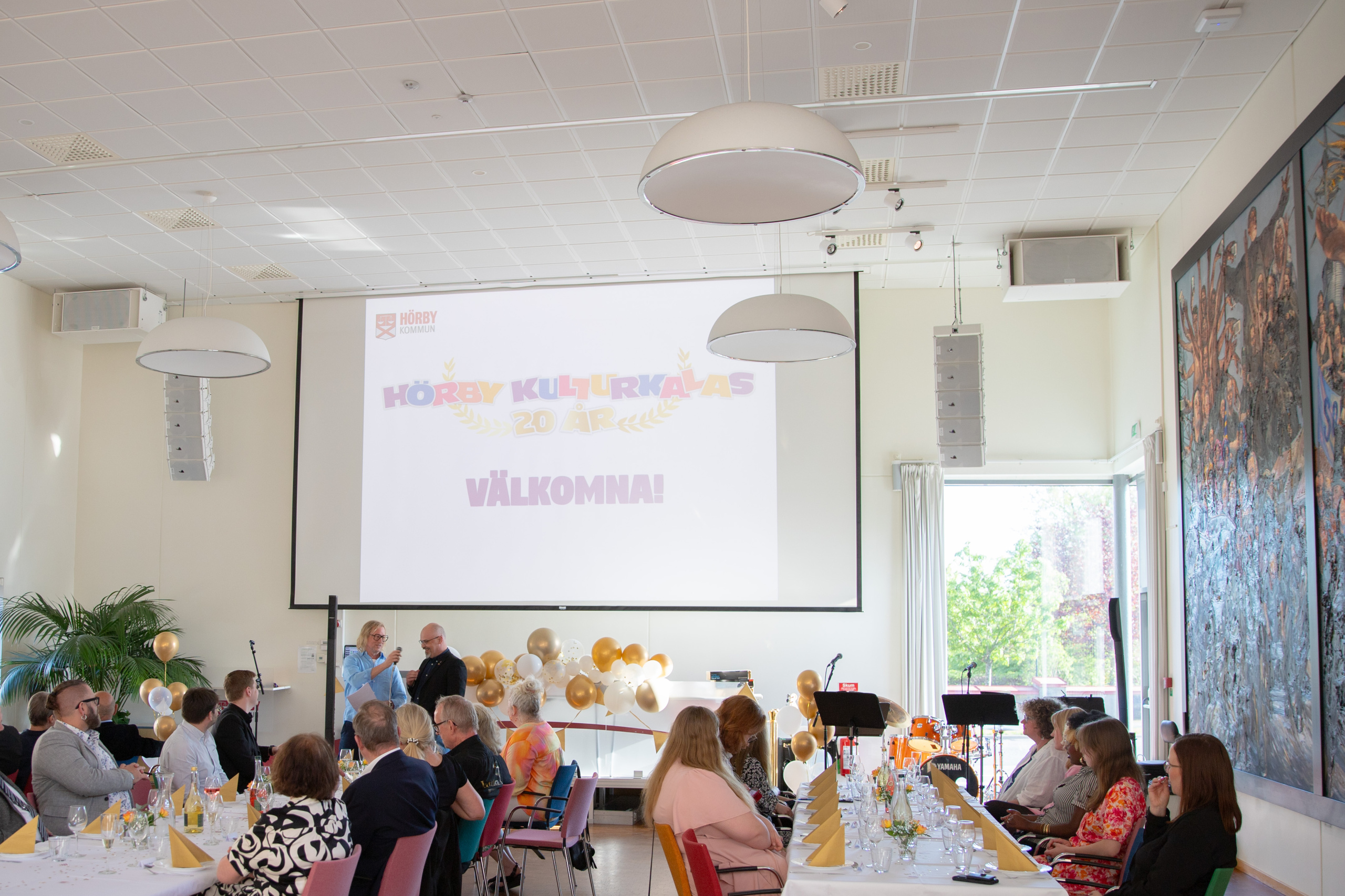 uppdukade långbord fullt med folk. Längst fram står två personer och håller tal. En projektor projicerar kulturkalas-logotypen