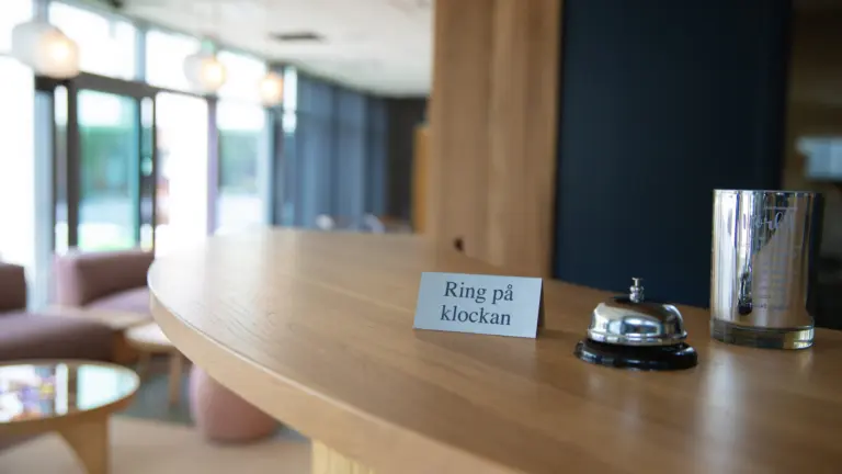 Närbild på receptionsdisk med ringklocka och skylt med texten "ring på klockan" i fokus.