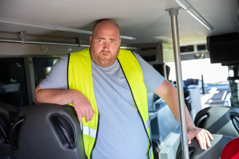 En man i gul varselväst står i en buss, lutar sig mot ett säte och tittar in i kameran.