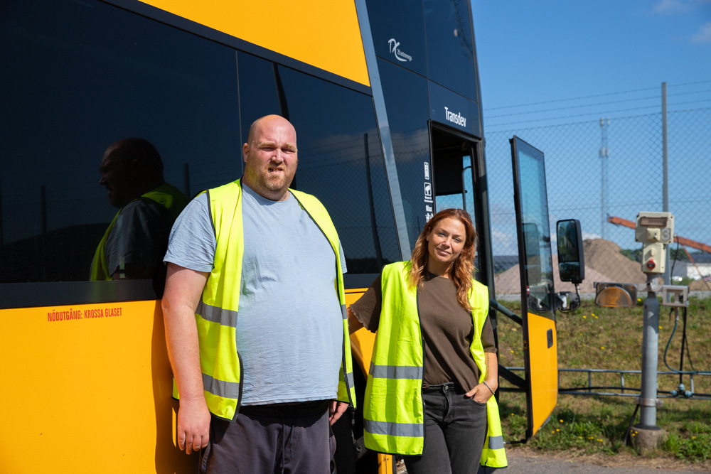 En man och en kvinna iförda varselvästar står framför en gul buss i solljus