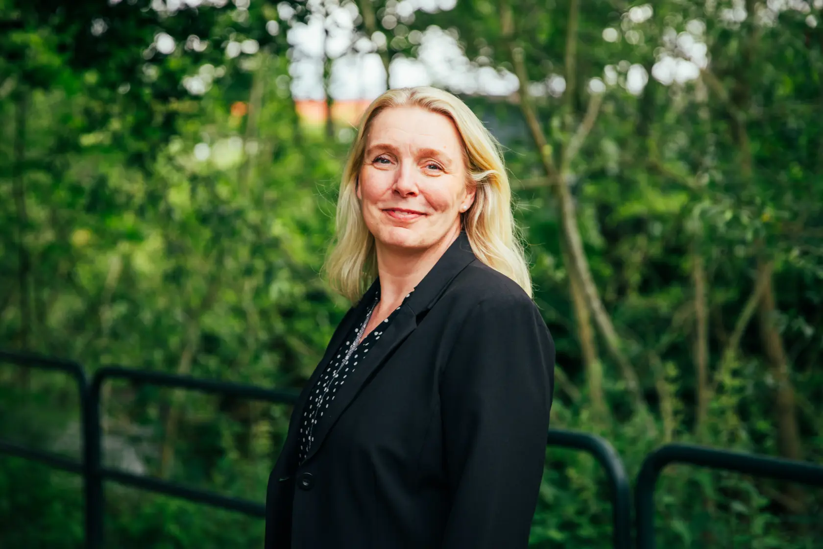 Halvbild på en blond kvinna som står bland grönska och ler in i kameran