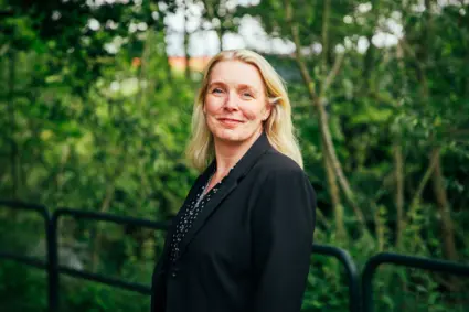 Halvbild på en blond kvinna som står bland grönska och ler in i kameran