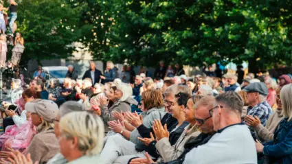 En stor publik siter på stolar utomhus och kollar på ett framträdande. Flera personer klapper händerna.