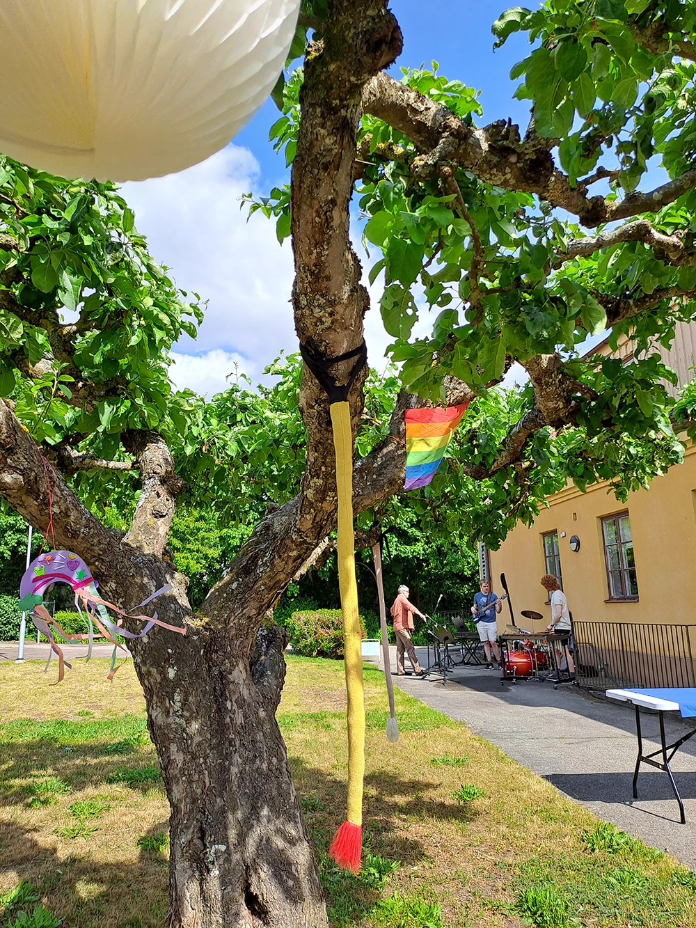 En bild på ett träd med flaggor och vindspel som står utanför en gul byggnad. Det är soligt ute. I bakgrunden syns musikanter som riggar upp sina instrument.