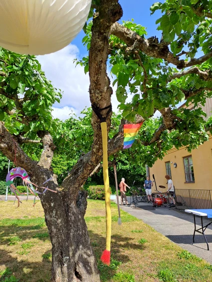 En bild på ett träd med flaggor och vindspel som står utanför en gul byggnad. Det är soligt ute. I bakgrunden syns musikanter som riggar upp sina instrument.