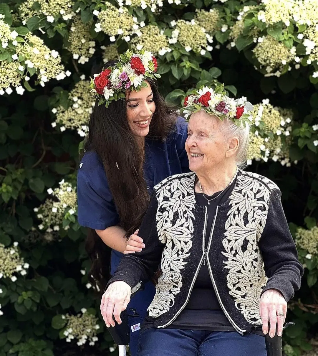 Ung kvinna som har armen om en äldre kvinna i rullstol, de har blommor i håret