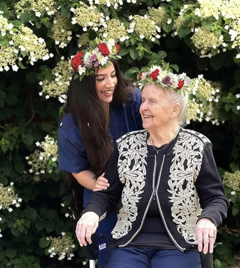 Ung kvinna som har armen om en äldre kvinna i rullstol, de har blommor i håret